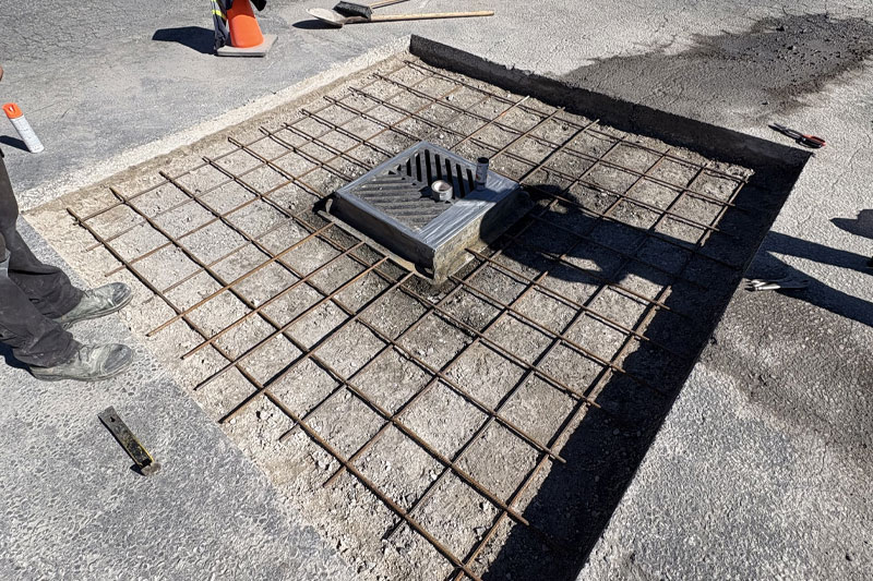 A square section of asphalt cut out around a storm drain, showing a metal reinforcement grid (rebar) laid over a gravel base in preparation for new concrete pouring. Allsite Services.