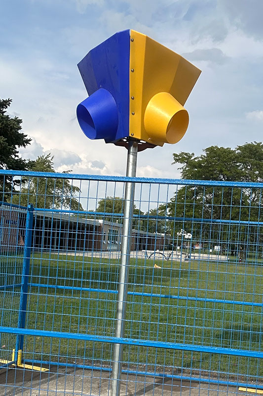 A blue and yellow multi-directional funnel ball hoop mounted on a tall silver pole in an outdoor playground setting, enclosed by a blue safety fence. Allsite Services.