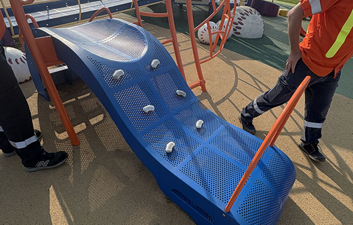Technicians performing safety maintenance and repairs on a blue textured playground climbing structure.