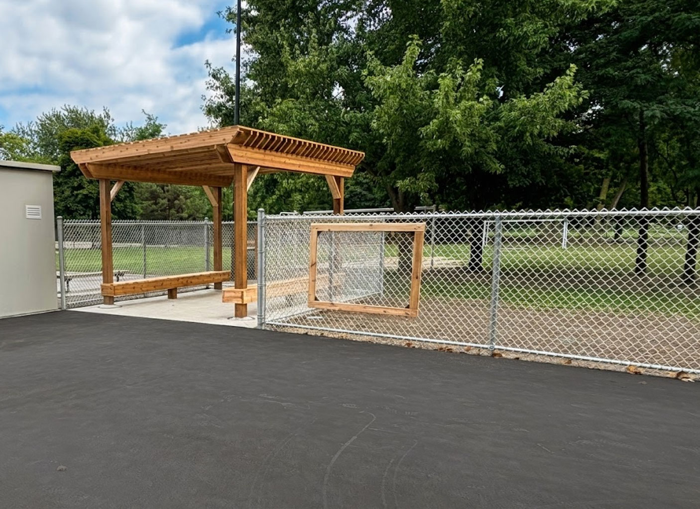 A heavy-duty timber frame pergola with a slatted roof providing shade over a concrete seating area. The structure is situated next to a chain-link fence bordering a grassy park with trees under a blue sky. Allsite Services.